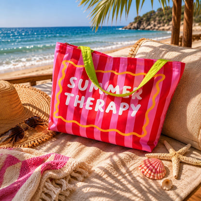Плажна чанта „Summer Therapy“ в розово и червено, поставена върху светъл плажен текстил до морето, със слънчеви отблясъци, мидички и летни аксесоари в уютна морска атмосфера.